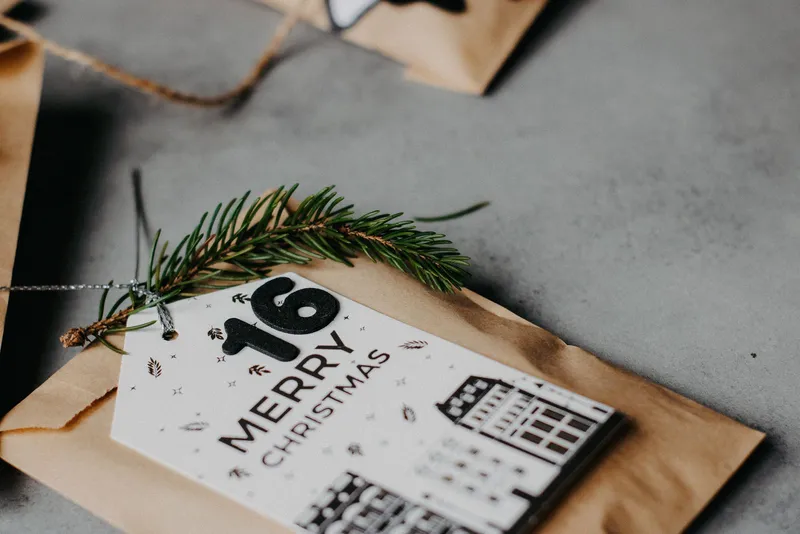 Sachet en papier kraft portant une étiquette de Noël numérotée 16 et un brin de sapin, sur un plan de travail gris