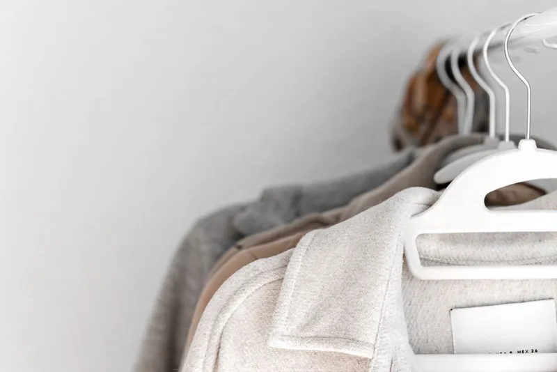 Vestes en laine écrue, beige et grise sur des cintres blancs, alignées sur une tringle contre un mur clair