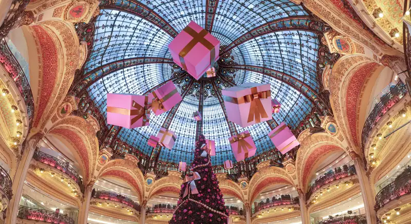 Sous une coupole de verre et de vitraux, de grands paquets roses noués de rubans dorés suspendus au-dessus d’un sapin sombre orné de boules roses, entre des balcons aux arcades dorées