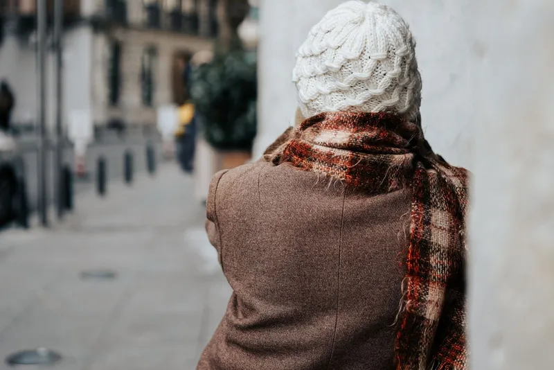 Femme vue de dos dans une rue, bonnet blanc en grosse maille torsadée et écharpe à carreaux rouille aux fils duveteux sur un manteau brun