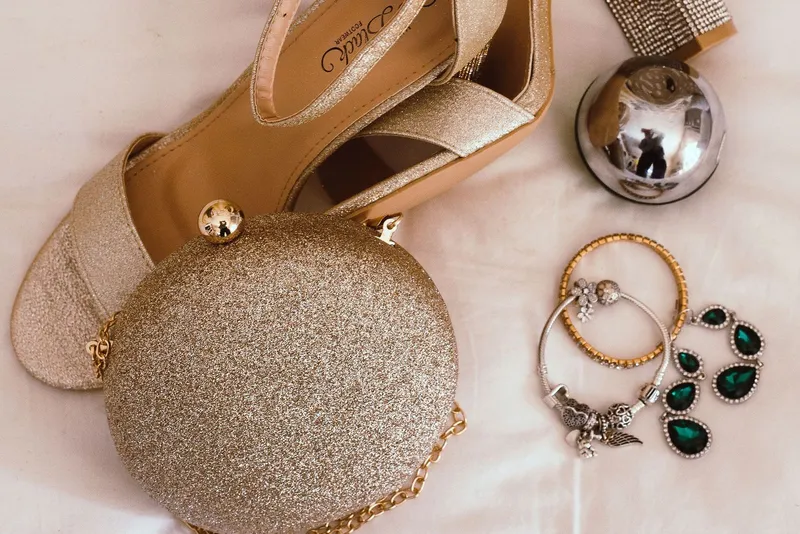 Sandales à talon pailletées dorées, pochette ronde scintillante à chaîne, bracelets et pendants d'oreilles à pierres vertes posés sur un drap blanc