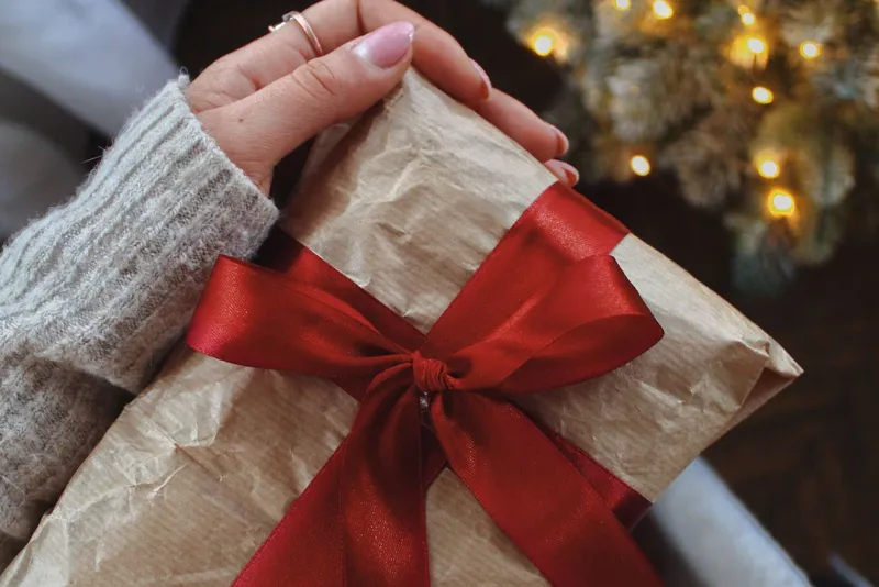 Main aux ongles roses tenant un paquet en papier kraft noué d'un ruban de satin rouge, près d'un sapin illuminé