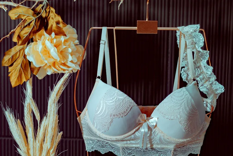 Soutien-gorge en dentelle ivoire à petit nœud de satin, suspendu à un cintre en forme de buste doré, près d'une pivoine et d'épis séchés