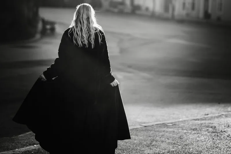 Photo en noir et blanc d'une femme aux longs cheveux clairs, vue de dos, qui soulève les pans d'un long manteau noir dans une rue