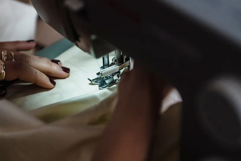 Mains aux ongles bordeaux guidant un tissu écru sous le pied-de-biche d'une machine à coudre