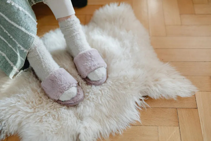Pieds en chaussettes duveteuses glissés dans des mules en fausse fourrure mauve, posés sur une peau de mouton blanche et un parquet en chevrons