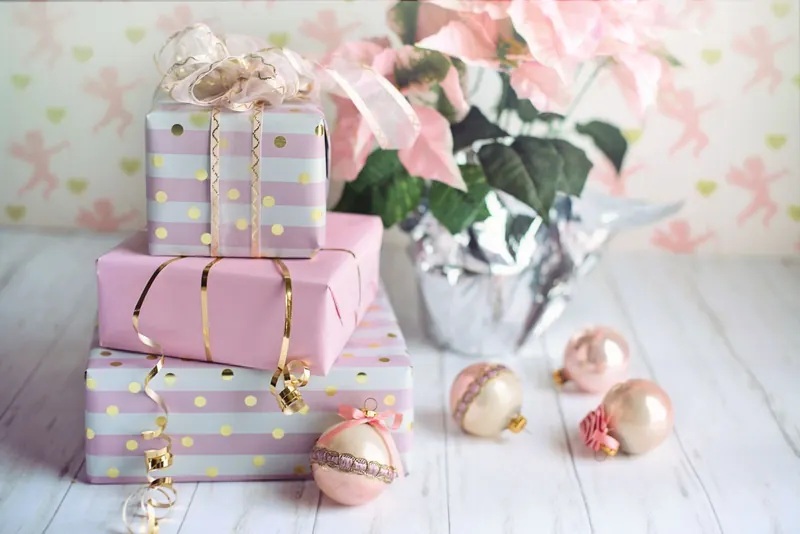 Trois paquets empilés, emballés de papier rose et de papier rayé rose et blanc à pois dorés, noués de rubans dorés, près d’un poinsettia rose et de boules de Noël nacrées
