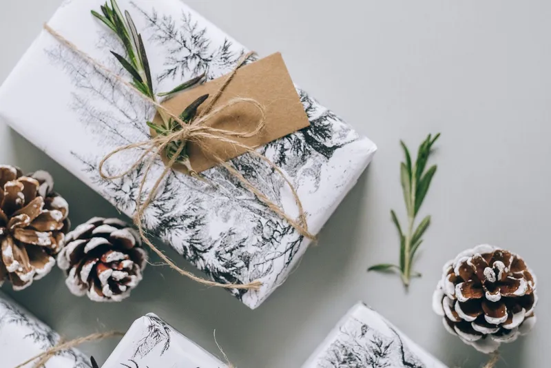 Petits paquets en papier blanc illustré de sapins, noués de ficelle avec une étiquette kraft et du romarin, entourés de pommes de pin