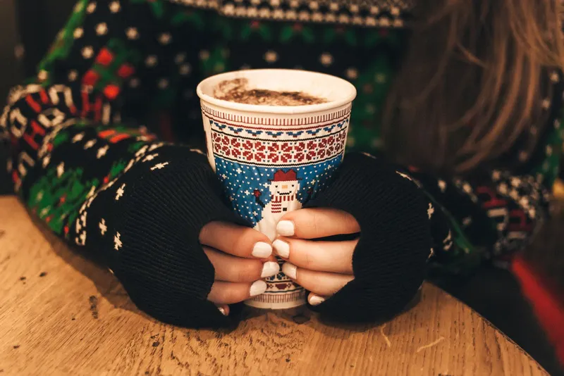 Mains en mitaines noires serrant un gobelet de chocolat chaud à motif de bonhomme de neige, devant un pull de Noël en jacquard vert, rouge et blanc