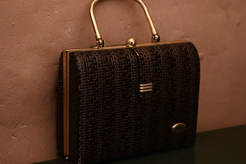 Sac à main rétro rigide à motif tressé brun, avec anse et fermoir en métal doré, posé contre un mur ocre rosé
