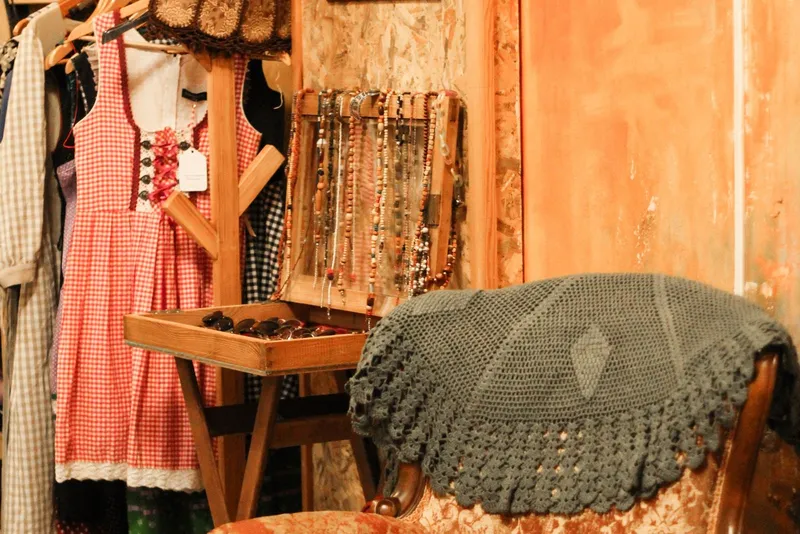 Coin de friperie avec une robe à carreaux rouges sur un portant, des colliers de perles en bois et un fauteuil ancien couvert d'un châle au crochet